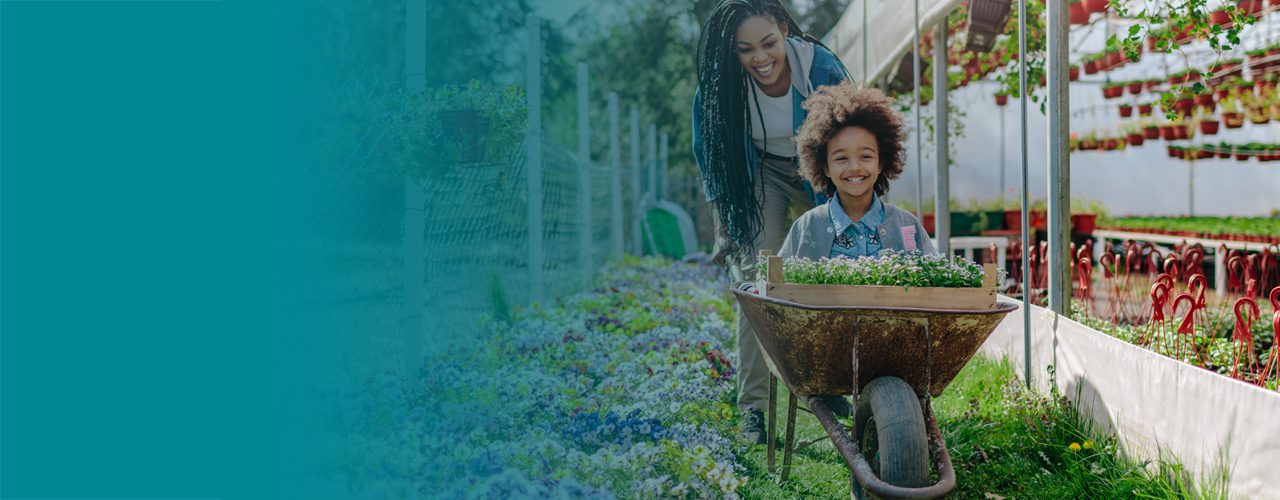 A smiling child sitting in a wheelbarrow filled with flowers, pushed by a joyful woman in a garden setting.